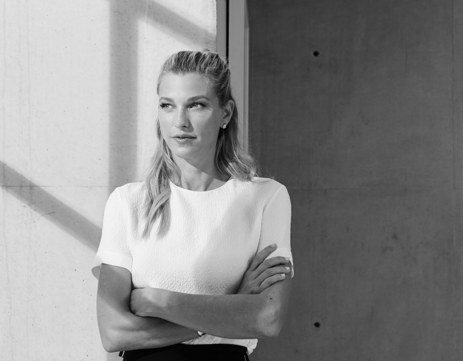 Nicole Junkerman steht in einem hellen Raum an einer Betonwand, die Arme locker verschränkt, und blickt ruhig zur Seite.